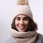 Person wearing a beige knit hat with a pom-pom and matching scarf against a white background